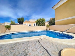 En swimmingpool med flisegulv, omgivet af en bygning med beige facade og en klar blå himmel.