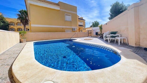 A swimming pool with a tiled edge, surrounded by a beige wall and a building with balconies in the background. - Pleasant 4 Bedroom Townhouse with Com Pool VM7 (Orihuela)