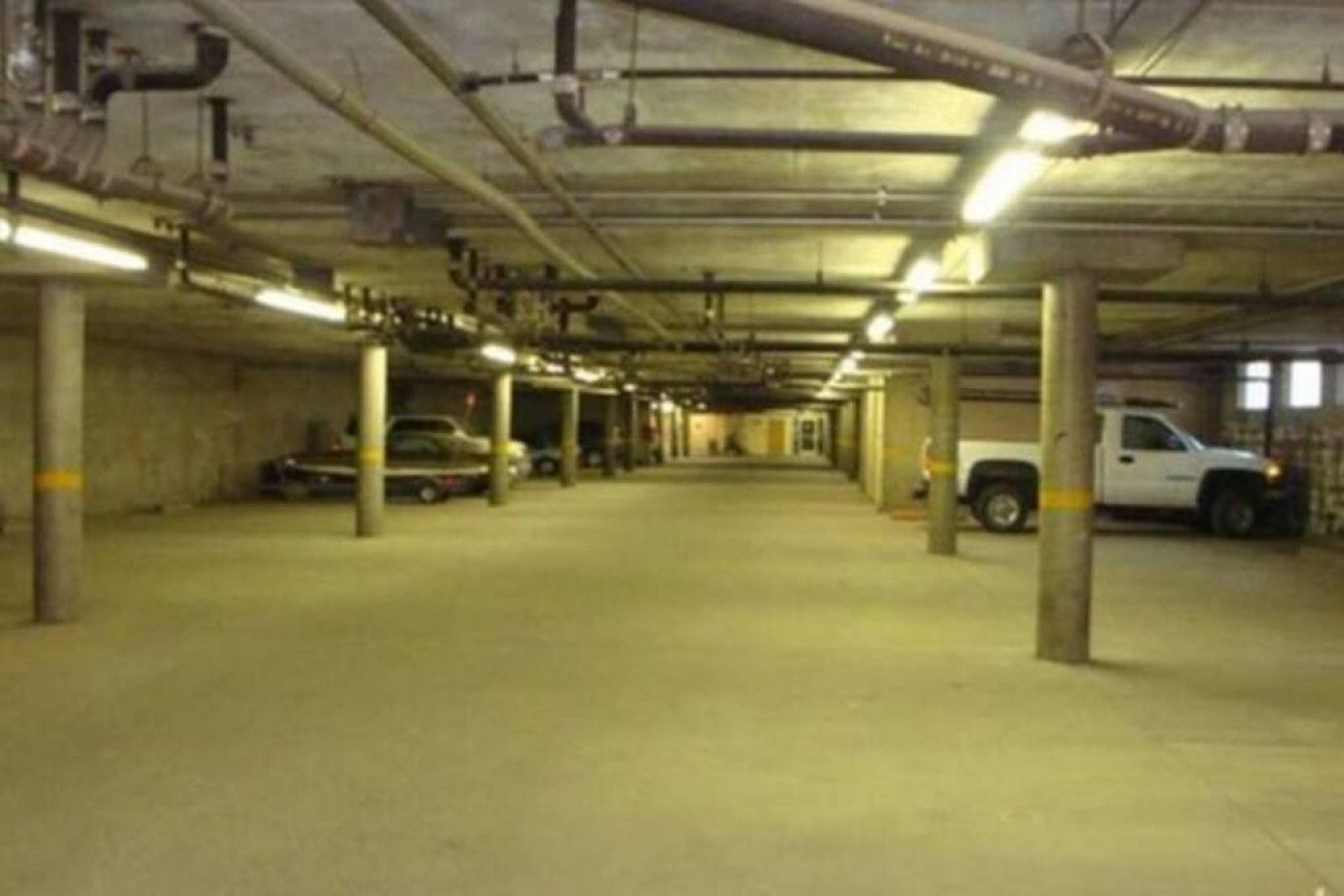 An underground parking garage with concrete pillars, fluorescent lighting, and parked vehicles.
