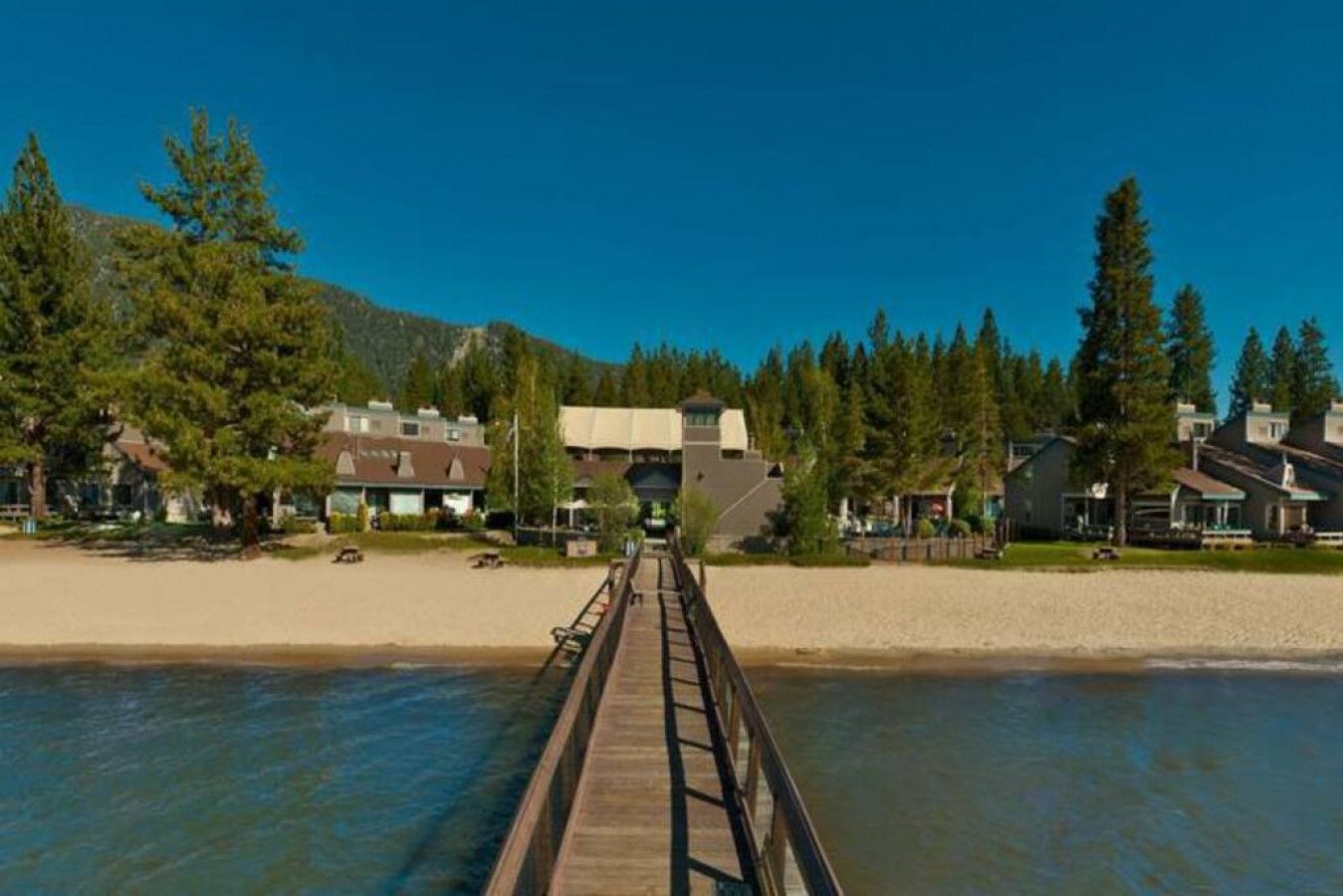 A wooden boardwalk leading to a building with a large overhang, surrounded by trees and a clear sky.