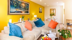 Un salón con sofá color beige, cojines azules y naranjas, una mesa de centro blanca con fruta, y un cuadro en la pared amarilla.
