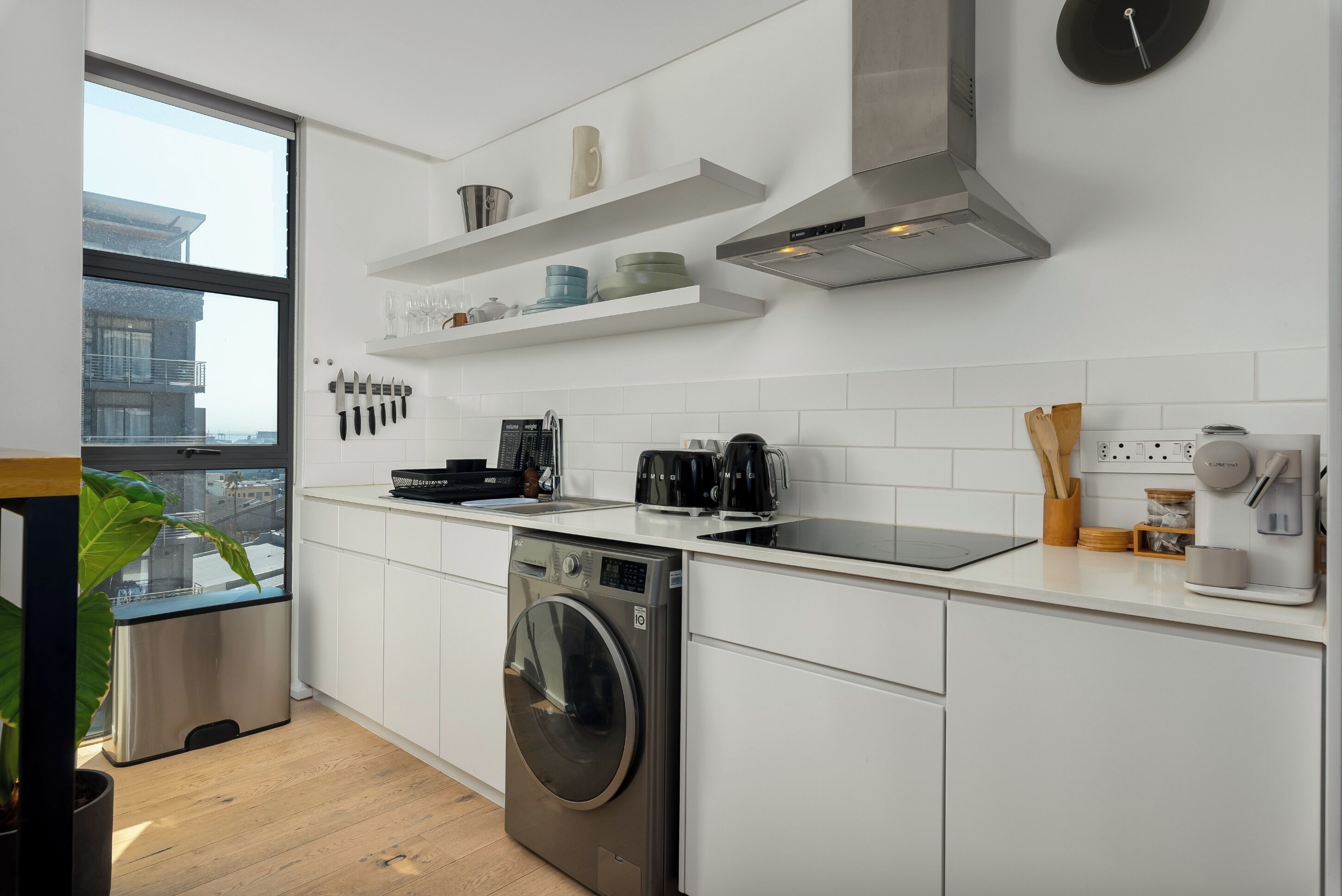 Une cuisine moderne avec des meubles blancs, un lave-linge encastré, une machine à café et une fenêtre donnant sur des immeubles.