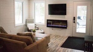 Een woonkamer met een flatscreen televisie boven een open haard, een bank, een fauteuil en een salontafel.