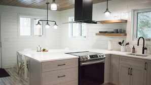 Een moderne keuken met een centraal kookeiland, een inbouwoven en roestvrijstalen apparatuur. Er hangen hanglampen boven het eiland en een raam zorgt voor natuurlijk licht.