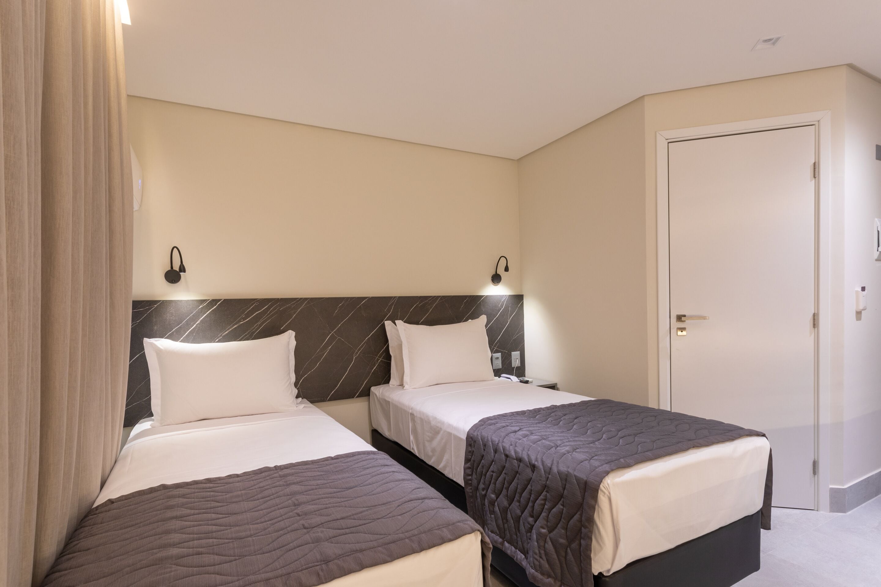 Habitación de hotel con dos camas, cabecera de mármol, una puerta y una mesita de noche con lámpara.