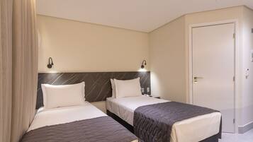 Quarto de hotel com duas camas, cabeceira de mármore, uma porta e mesa de cabeceira com abajur.