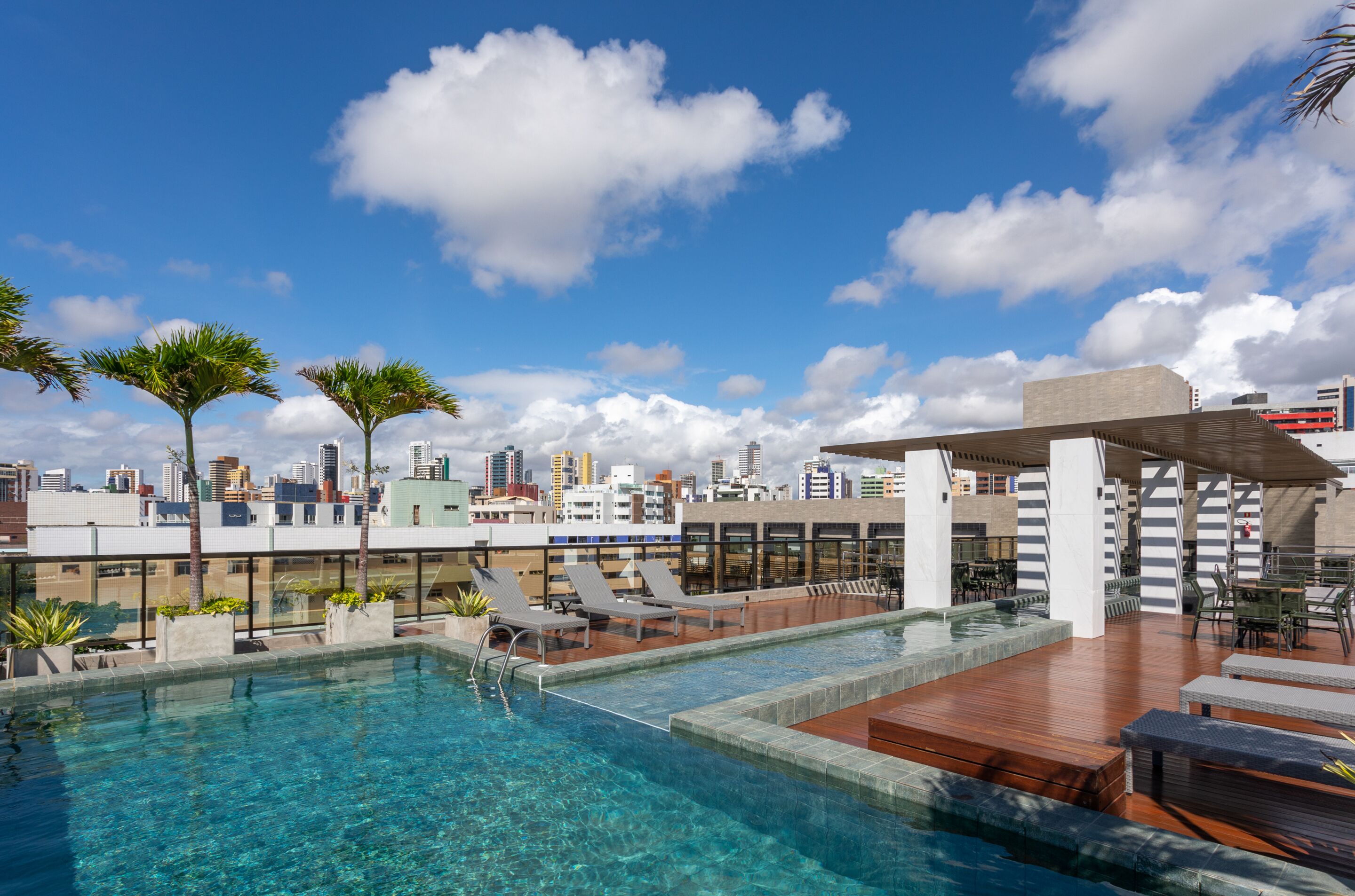 Una piscina en la azotea con agua azul cristalina, sillones de descanso y el perfil de la ciudad al fondo.