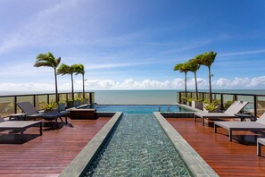 Piscina infinita no terraço com espreguiçadeiras e plantas em vasos, com vista para um oceano azul e límpido.