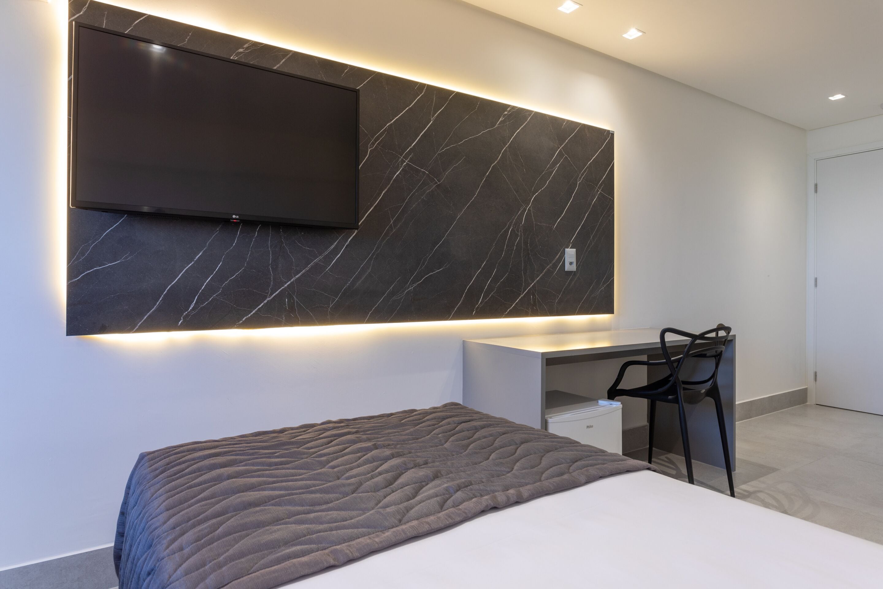 Un dormitorio moderno con una cama grande, un escritorio y un televisor montado en una pared de mármol.