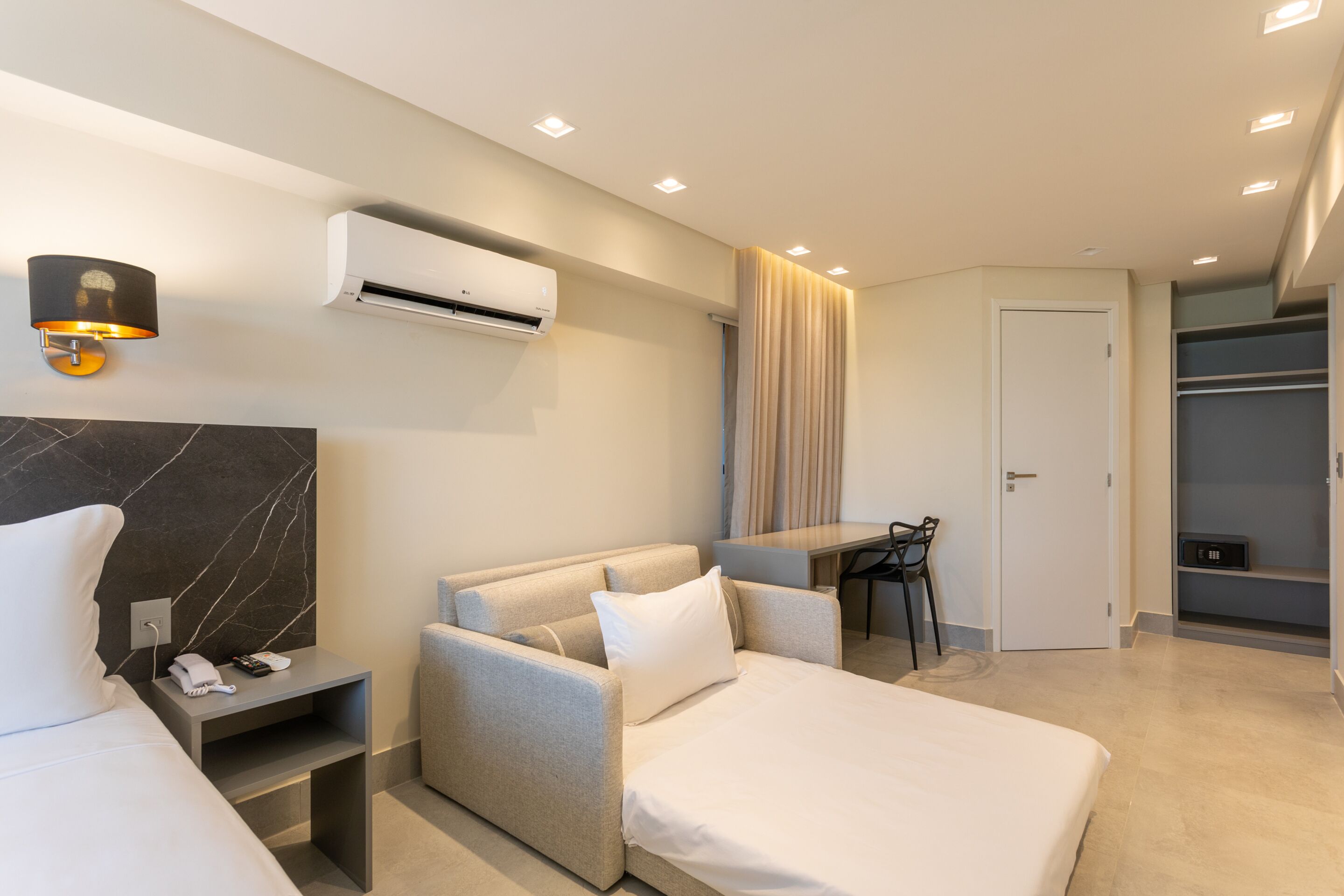Una habitación de hotel moderna con una cama, un sofá, un escritorio y una silla. Se observa una unidad de aire acondicionado en la pared y una lámpara en la mesita de noche.