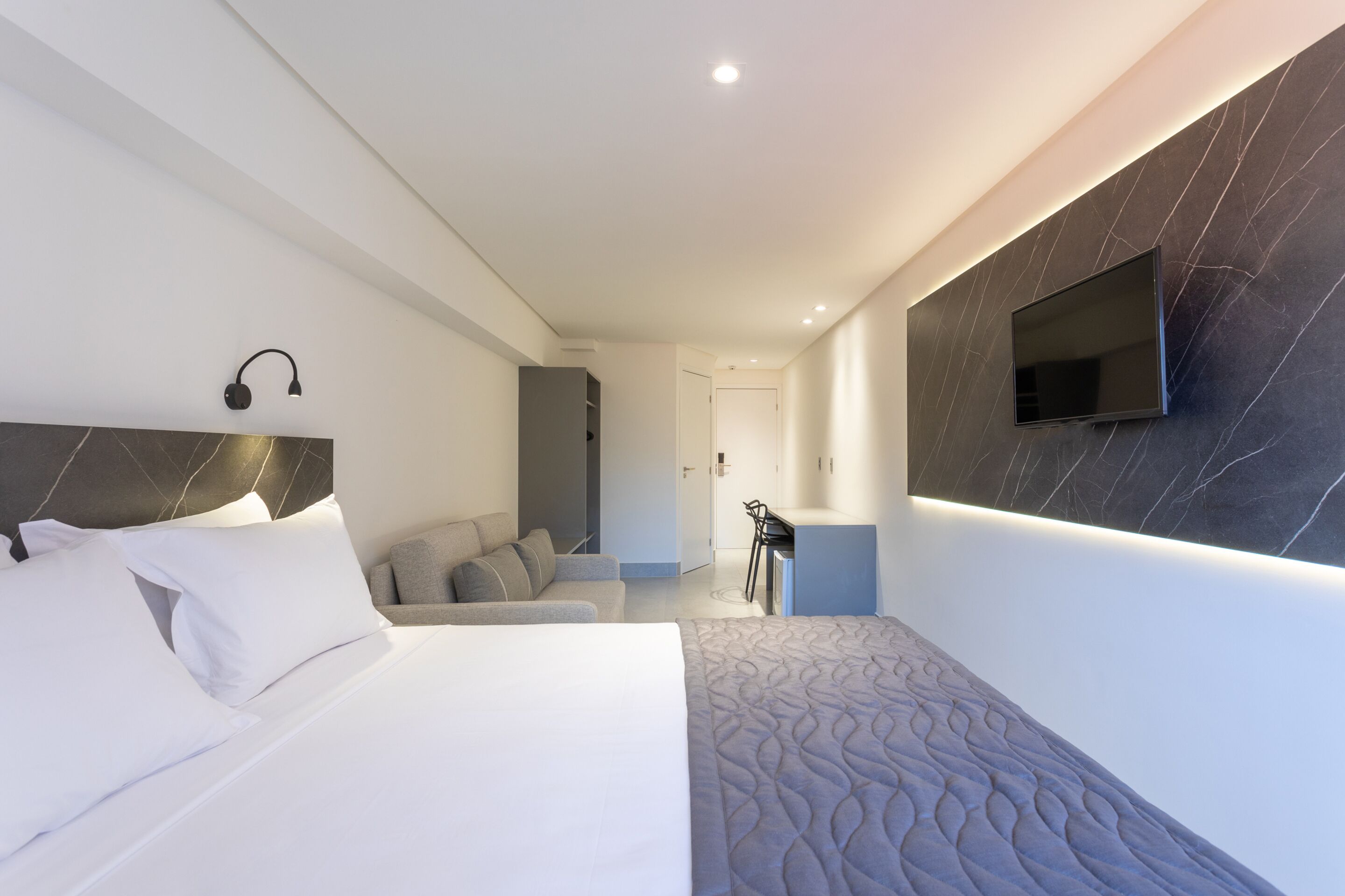 A modern hotel room with a large bed, a desk, a chair, and a TV mounted on the wall.