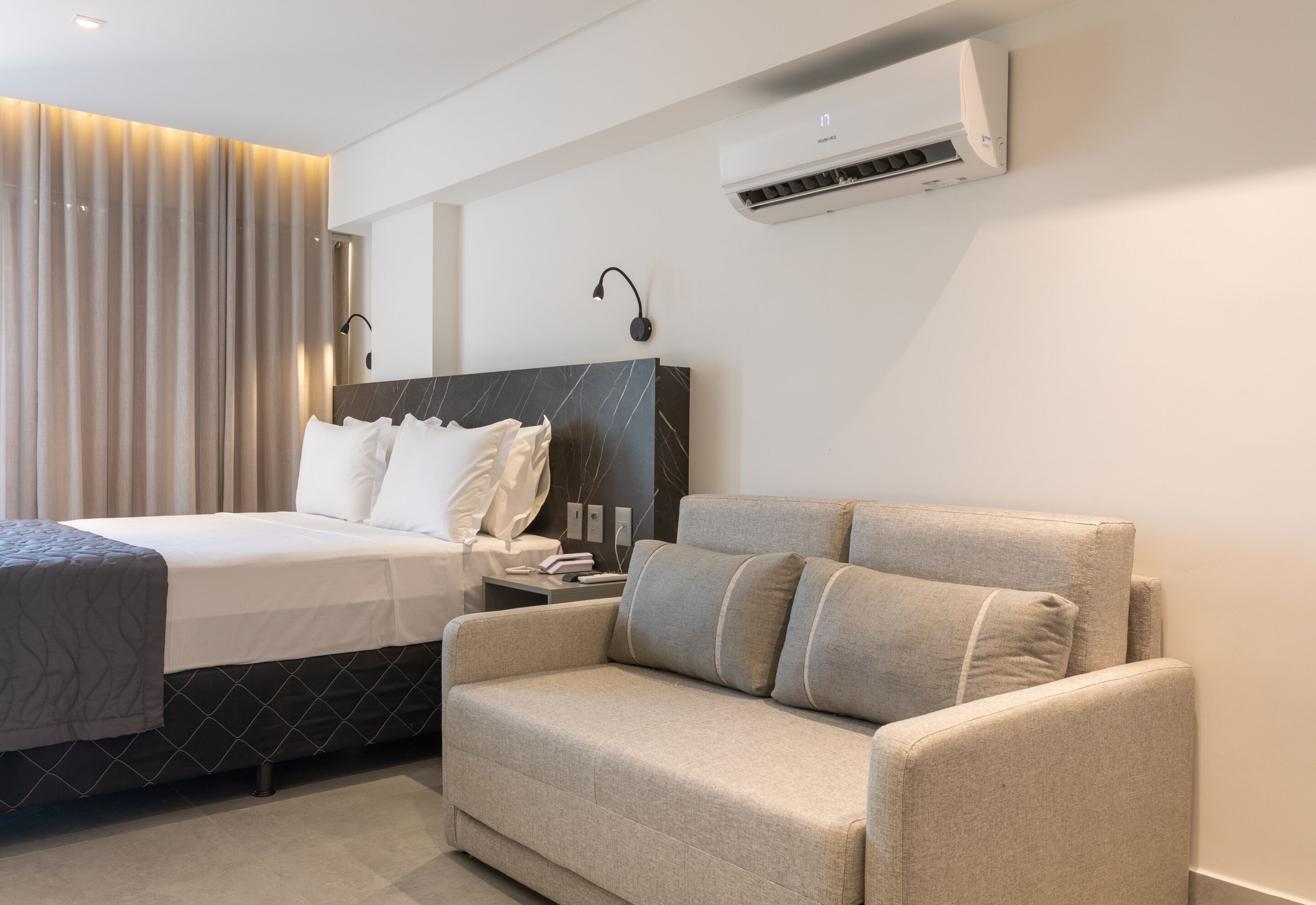 Una habitación de hotel moderna con cama, sofá, aire acondicionado y una lámpara fijada a la pared.