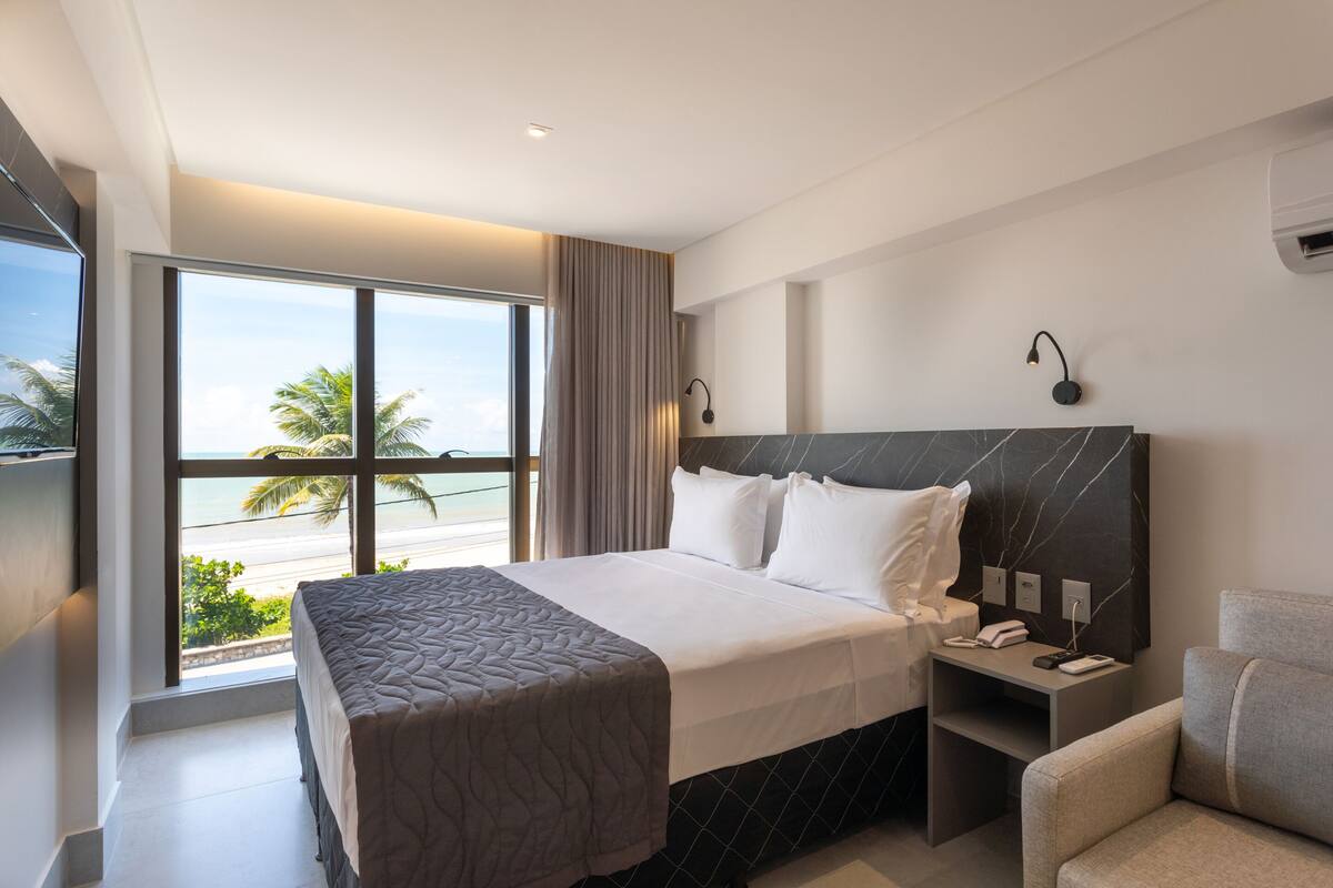 Una habitación de hotel moderna con una cama grande, una mesita de noche, una silla y vista a la playa.