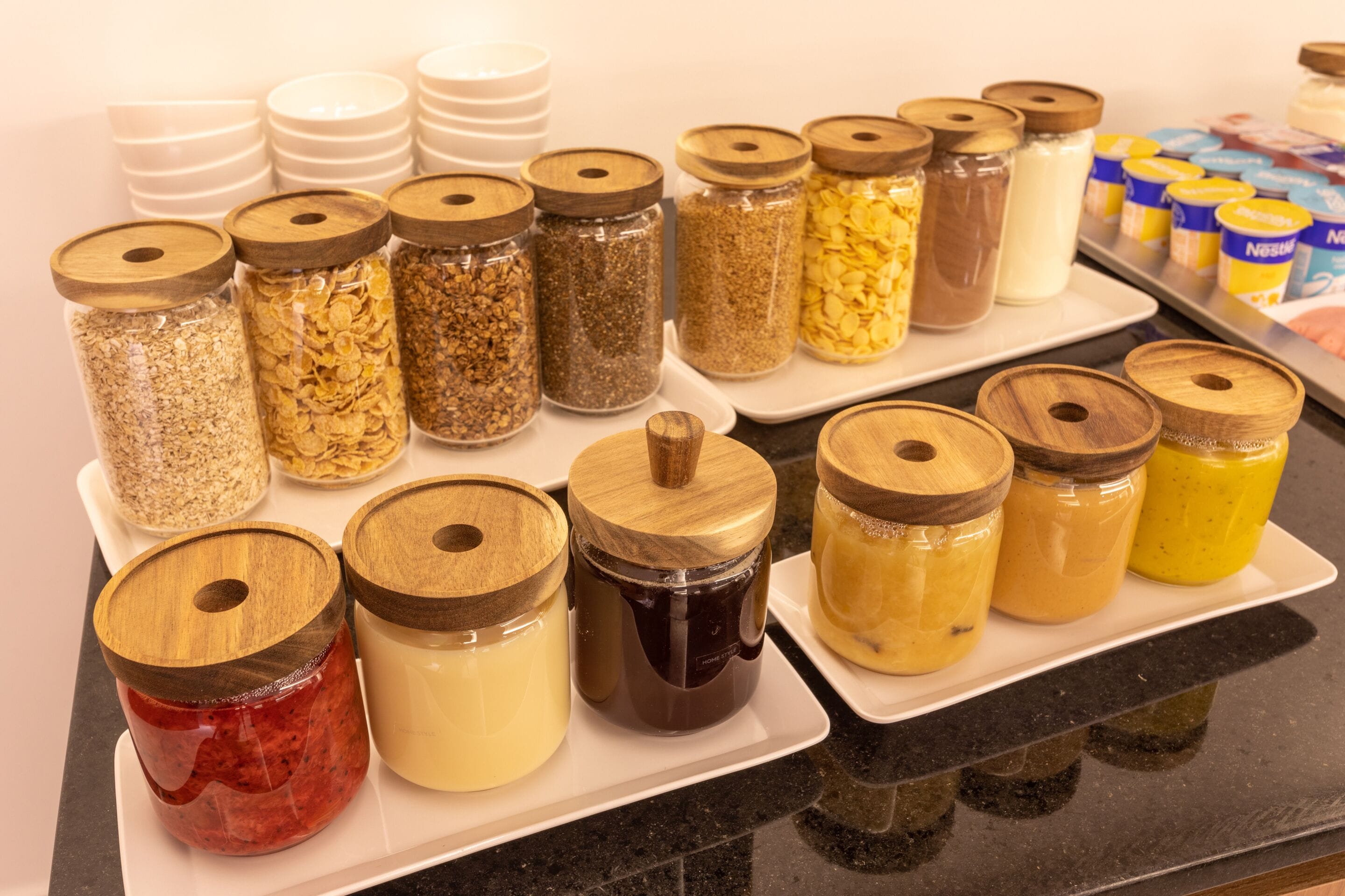 A variety of food items in jars with wooden lids, arranged on a white tray.