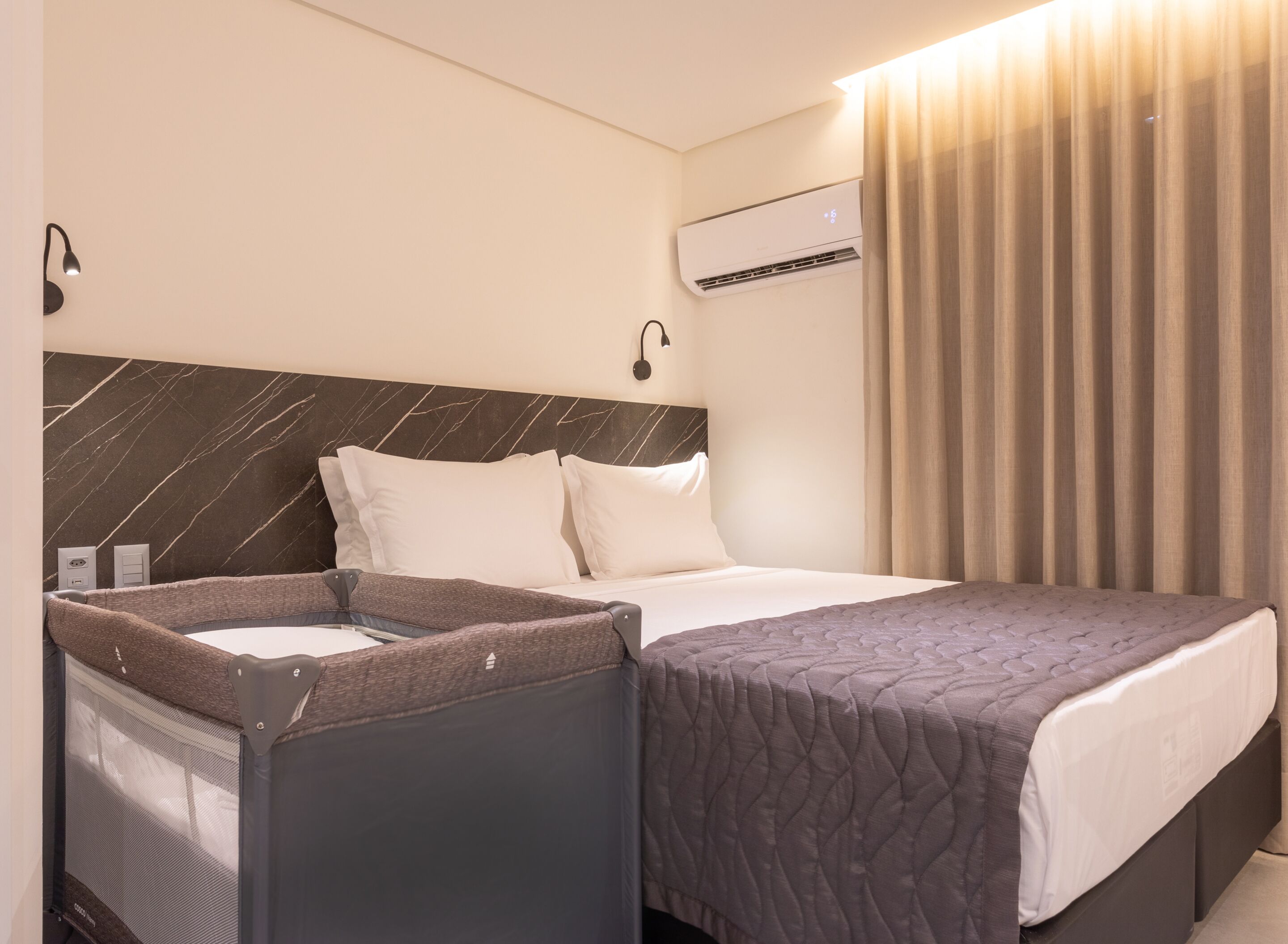 A hotel room with a bed, a small seating area with a screen, an air conditioning unit, and a curtain.