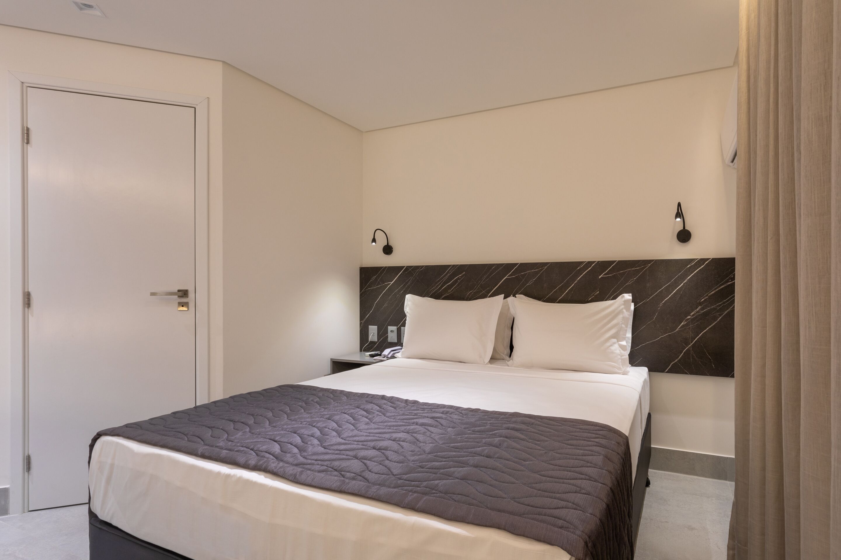 Habitación de hotel con cama, mesita de noche, lámpara de pared y una puerta.