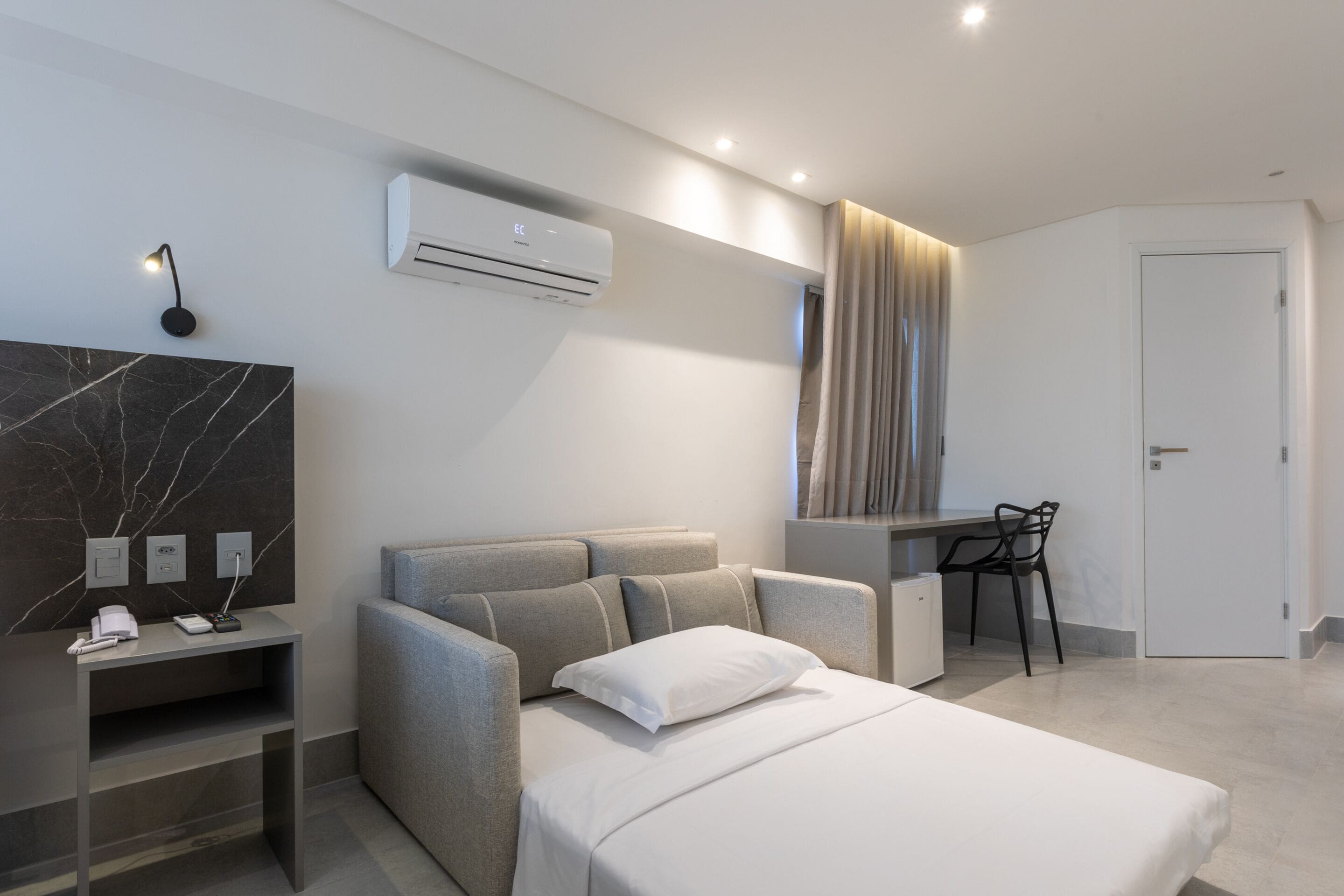 Una habitación de hotel moderna con cama, escritorio, silla, unidad de aire acondicionado y pared de mármol.