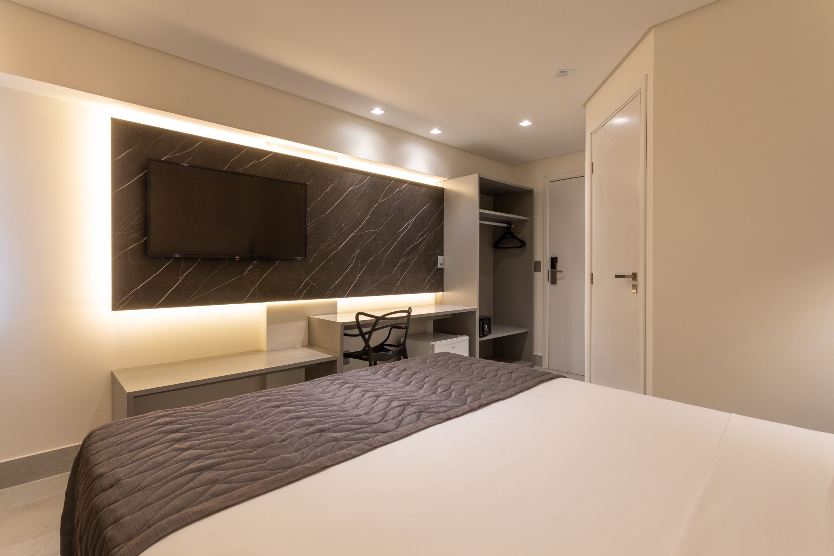 Un dormitorio moderno con una cama grande, un escritorio, una silla, un televisor montado en una pared de mármol y un armario.
