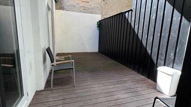 A wooden deck with a metal chair, a white trash bin, and a black railing. - The Old Factory - Hostel (Copenhagen)