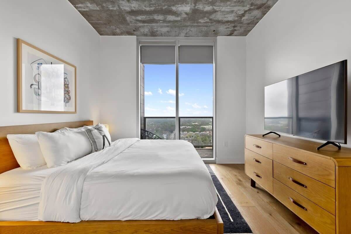 A modern bedroom with a large bed, a wooden dresser, a flat-screen TV, and a city view through the window.