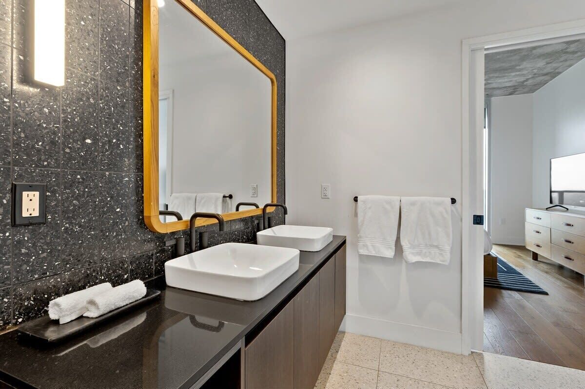 A modern bathroom with a black speckled backsplash, two white sinks, a large mirror with a gold frame, and a towel rack with white towels.