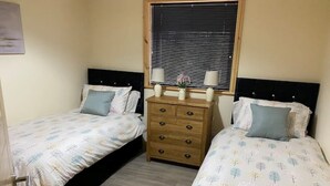 A bedroom with two beds, a wooden dresser, and a window with blinds.