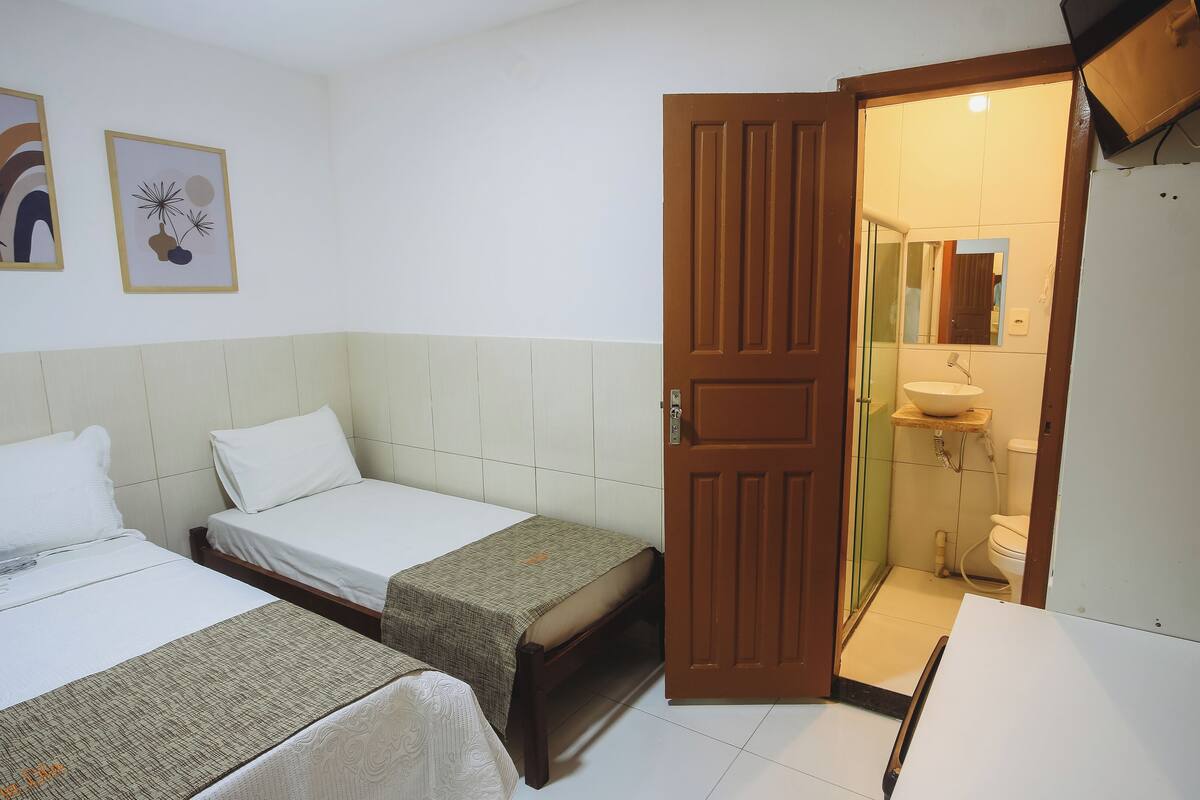 Habitación de hotel con dos camas individuales, una puerta de madera, baño con lavamanos e inodoro, y dos cuadros enmarcados en la pared.