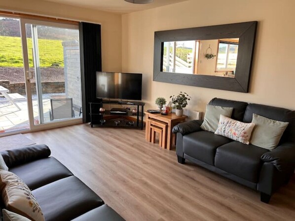 Ein Wohnzimmer mit einem schwarzen Ledersofa, einem Fernseher auf einem schwarzen Standfuß, einem kleinen Holztisch mit einer Pflanze und einem großen Fenster mit Blick auf ein grünes Feld.