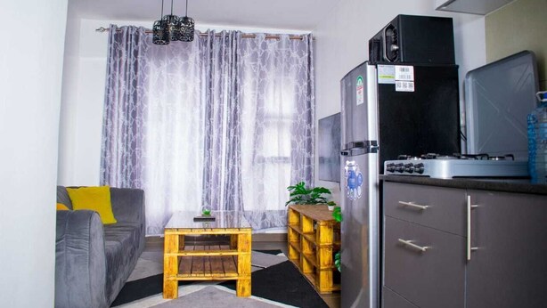 Um espaço de estar compacto com cozinha compacta, um sofá cinzento com almofadas amarelas, uma mesa de centro de madeira e uma janela grande com cortinas leves.