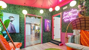 Una habitación con pared verde, un letrero de neón, una silla de mimbre y un sofá con cojines naranjas.