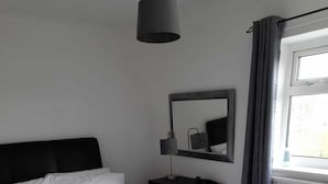 Un lit bien fait, avec du linge de lit blanc et des serviettes pliées, une table de chevet noire avec une lampe, un miroir et une fenêtre avec des rideaux.