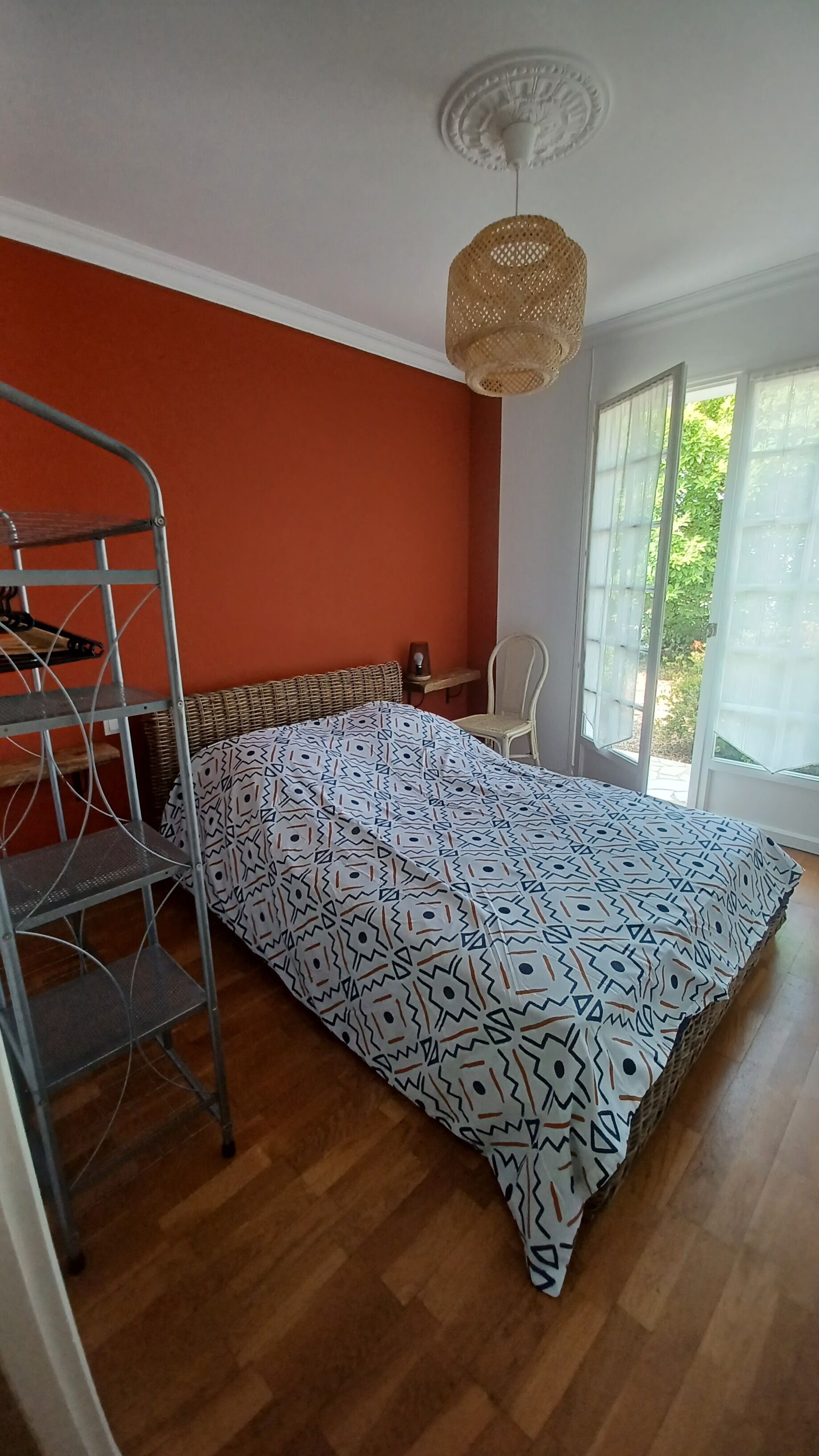 A room with a patterned bedspread, a metal shelf unit, a wicker chair, and a window with a view of greenery.