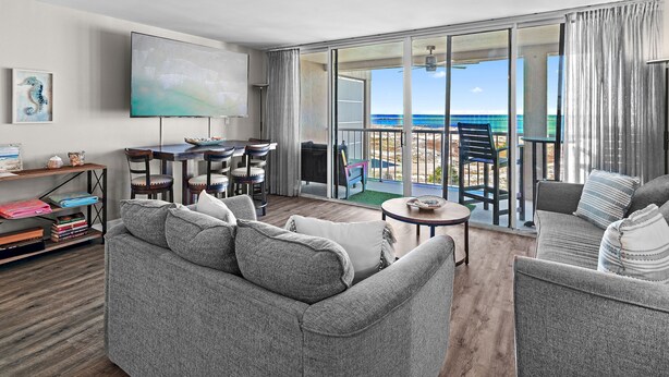 Un salon moderne avec un canapé gris, un téléviseur à écran plat et un coin repas comprenant une table et des chaises. On aperçoit un balcon offrant une vue sur l’océan.