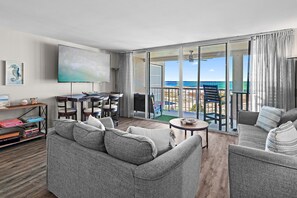 A modern living room with a grey sofa, a flat-screen TV, and a dining area with a table and chairs. A balcony with a view of the ocean is visible. - 1BR Beach Retreat with Balcony + Resort Amenities | Magnolia House 508 (Destin)