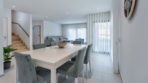 Una sala de estar moderna con una escalera, un comedor con una mesa blanca y sillas grises, y un reloj de pared grande.