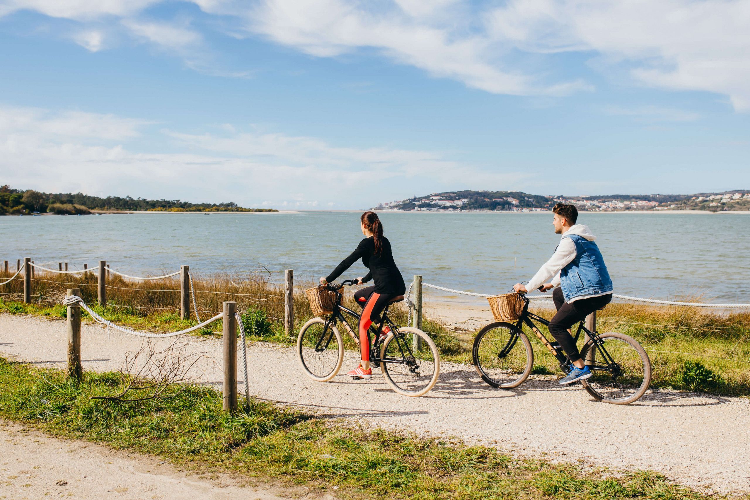 Two people riding bicycles on a sandy path by a body of water, with a rope barrier and wooden posts.