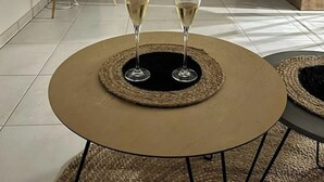 A round table with two wine glasses on a woven coaster, a television mounted on the wall, and a wooden cabinet in the background. - A haven of peace in the heart of the city (Reims)