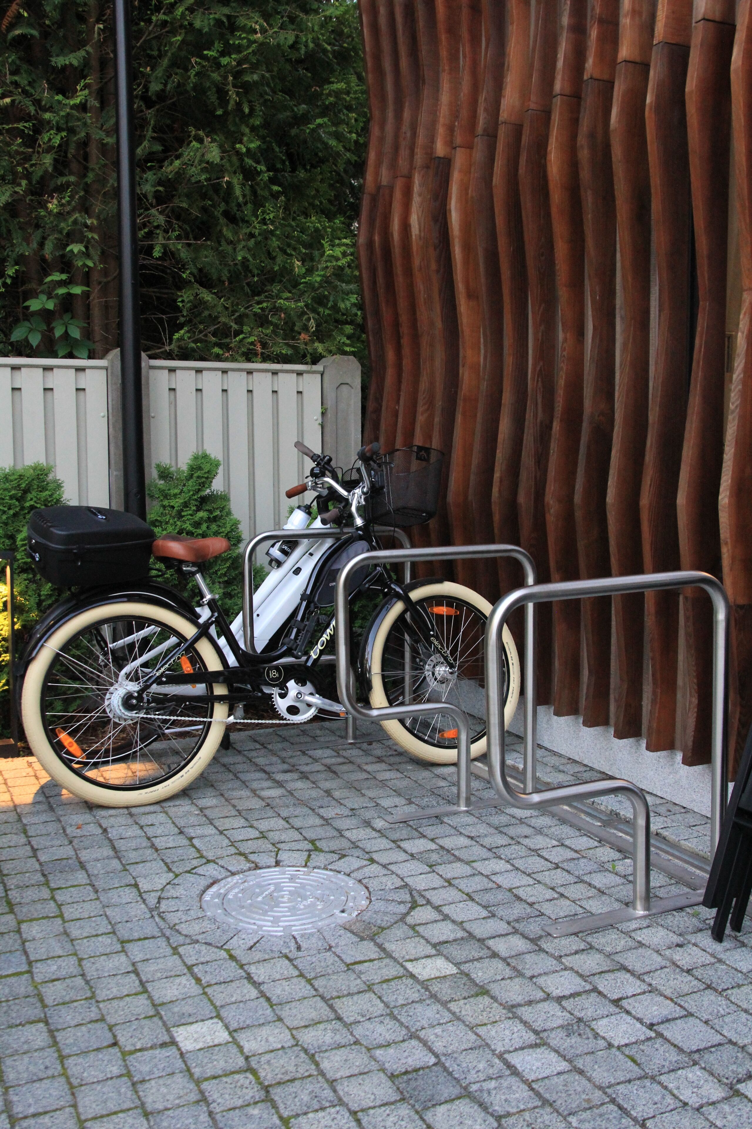 Una bicicleta estacionada al lado de un soporte metálico para bicicletas, con una valla blanca y follaje al fondo.