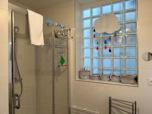 A shower with a glass door, a towel hanging on the door, and a window with a cloud decoration and hanging bags.