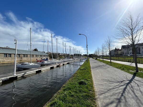 Um canal com barcos atracados à esquerda, um passeio pavimentado à direita e edifícios ao fundo.