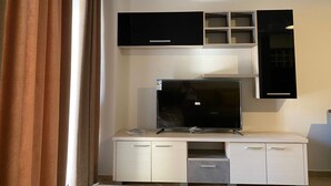 A modern living room with a flat-screen TV on a white cabinet, a black entertainment unit above, and a window with curtains. - Jey Vlora Apartaments (Vlorë)