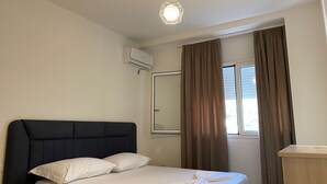 A bedroom with a bed, a bedside table, a window with curtains, and an air conditioning unit. - Jey Vlora Apartaments (Vlorë)