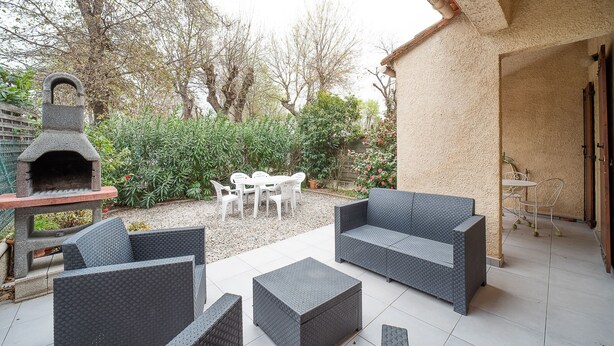 Un patio exterior con mobiliario de mimbre, una barbacoa de piedra y un jardín con árboles y plantas.