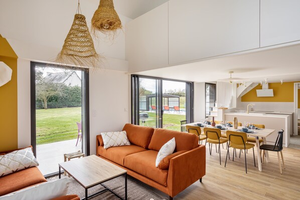 A modern living room with a sofa, a dining table, and chairs. There is a kitchen area with a sink and a window overlooking a garden.