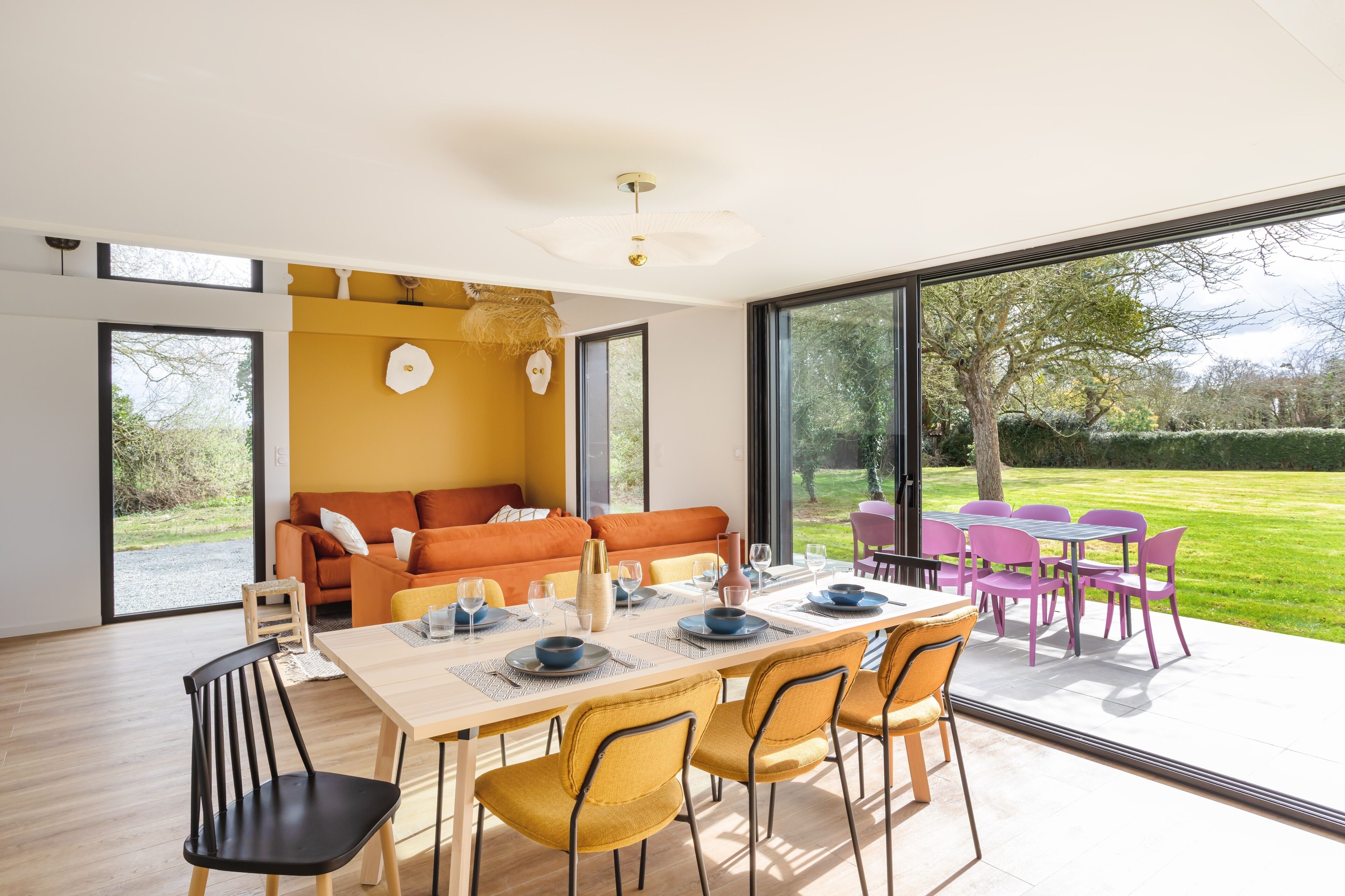 A modern dining area with a wooden table, yellow chairs, and a large window overlooking a garden.