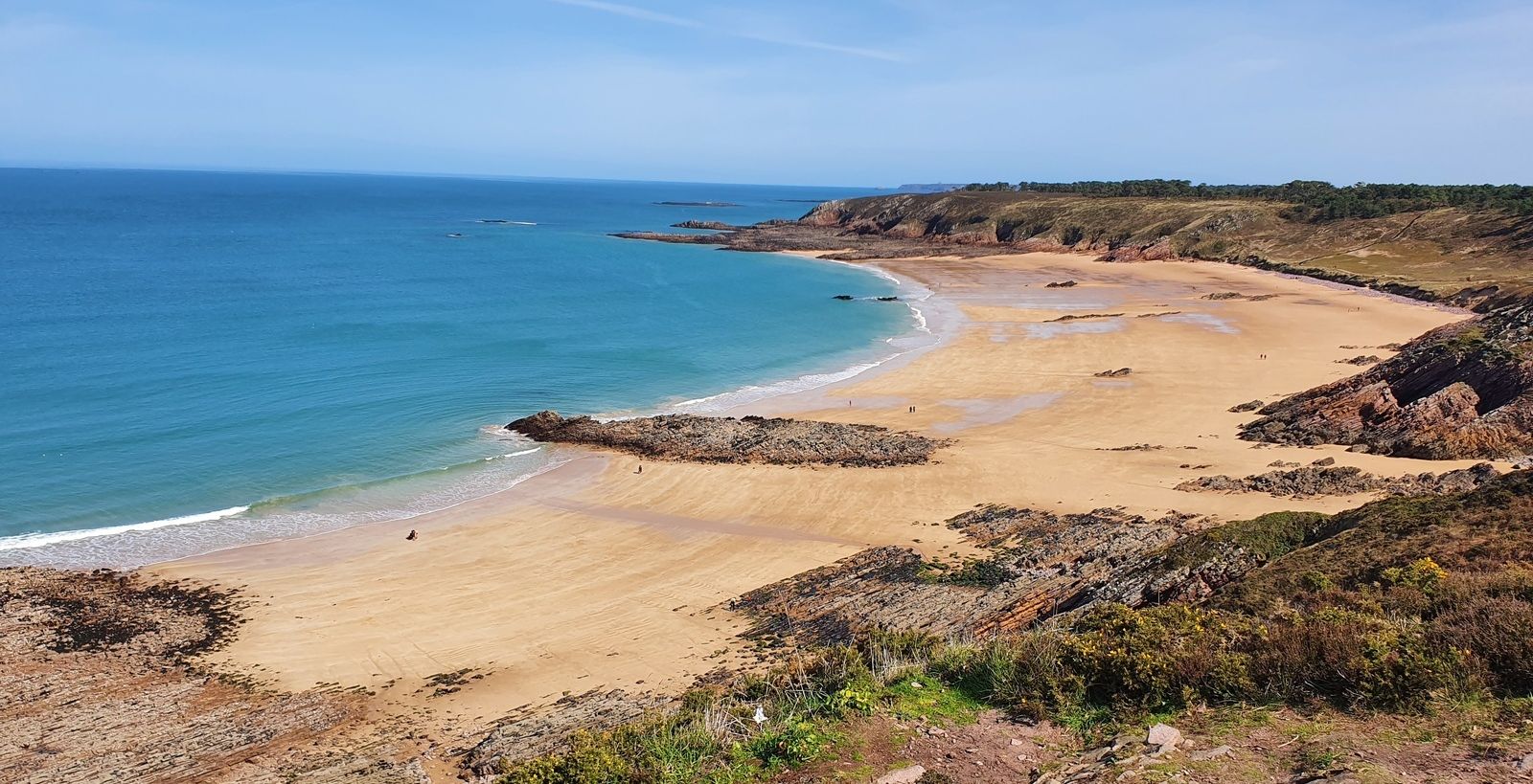 A coastal landscape with a sandy beach, rocky outcrops, and a clear blue sky.