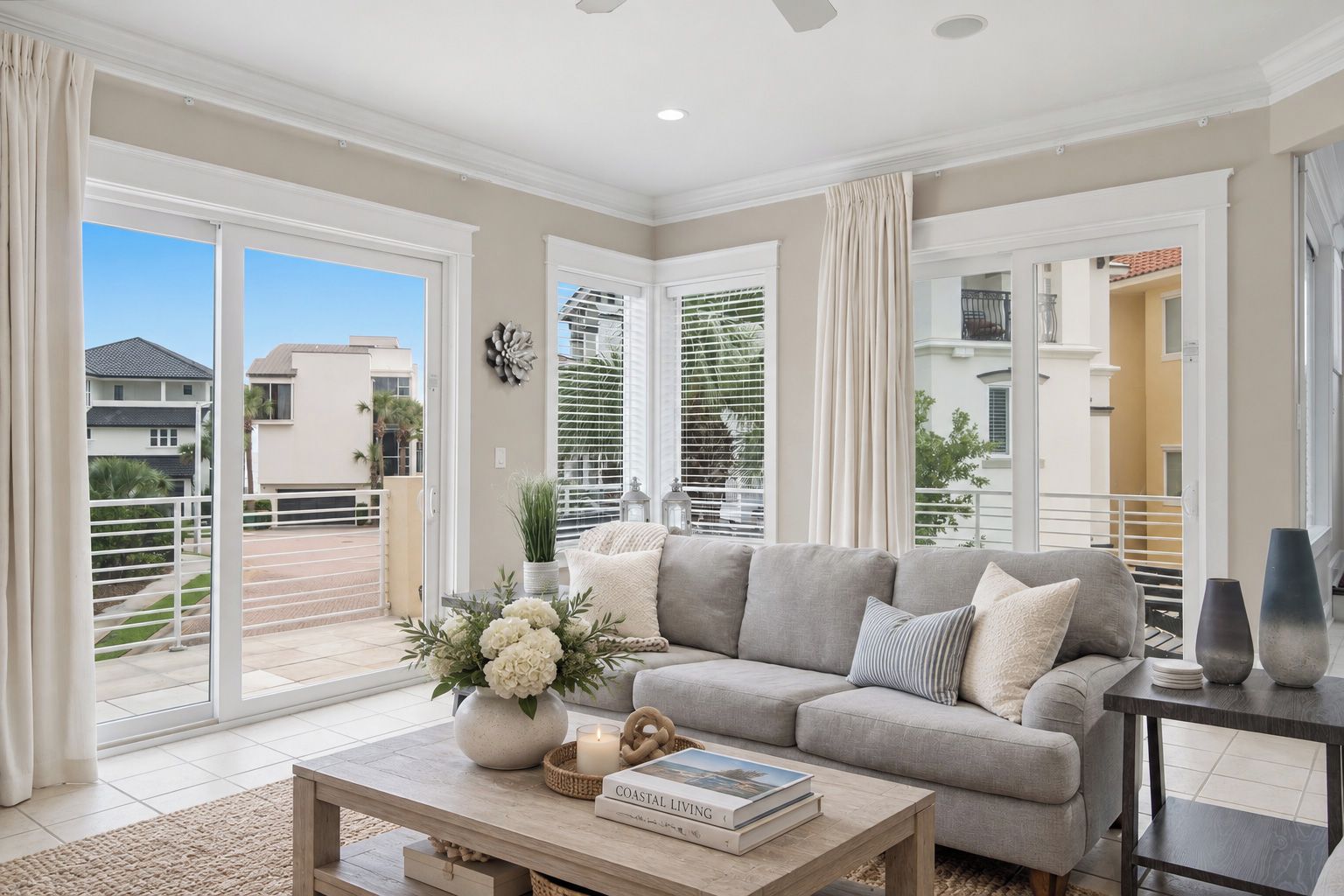 A living room with a sofa, a coffee table with a book and a candle, and a view of a residential area through large windows.
