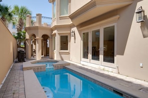 A swimming pool with a tiled deck, a covered patio area, and a building with large windows and a balcony.