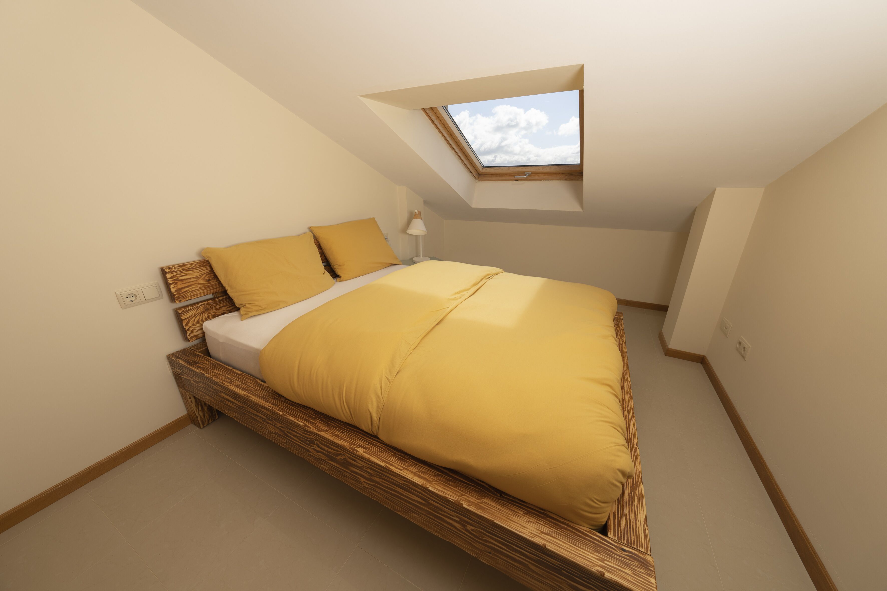 Un dormitorio con techo inclinado y tragaluces, con estructura de cama de madera y ropa de cama amarilla.