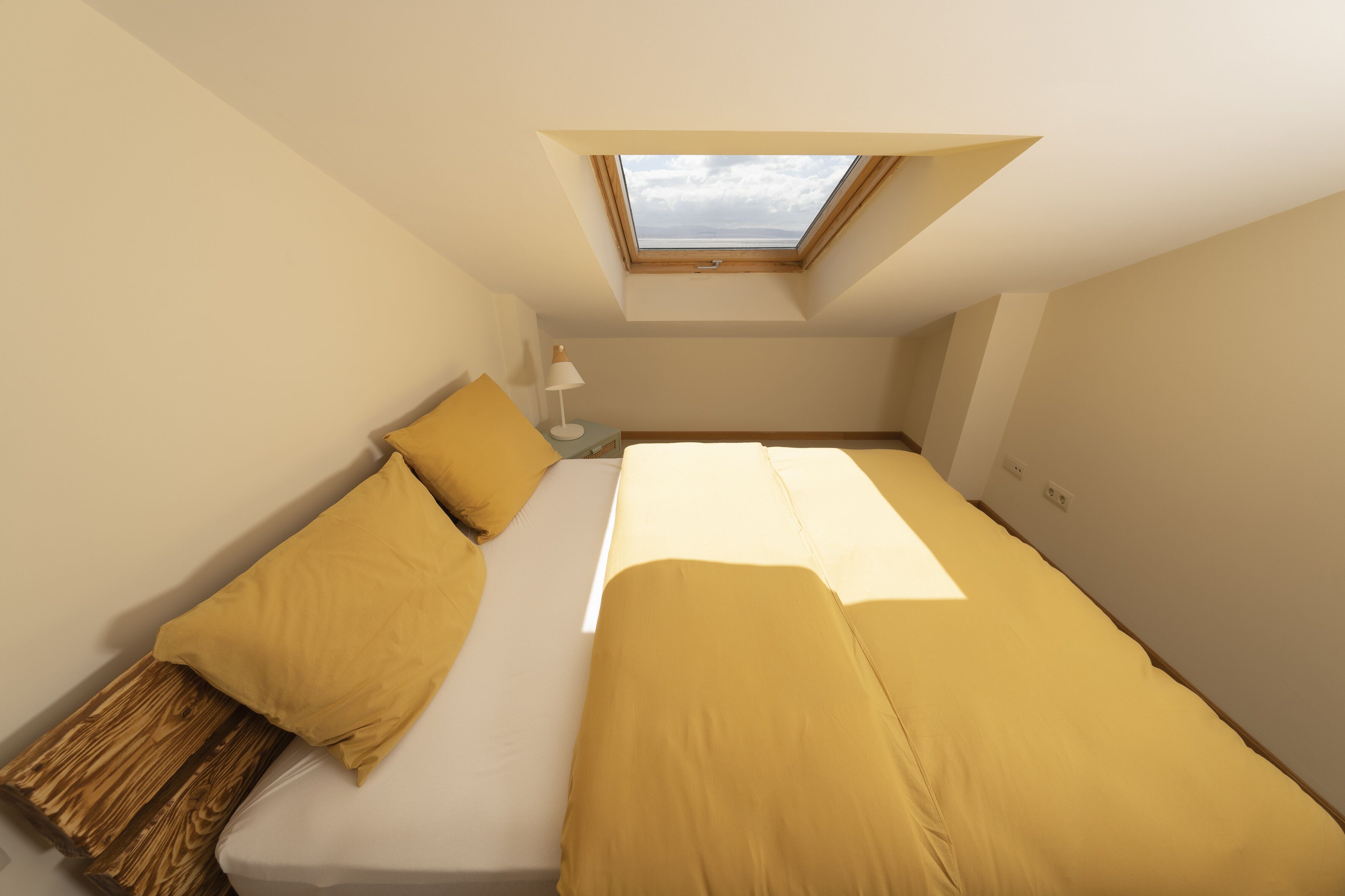 Una habitación pequeña con una cama individual, tragaluz, mesita de noche con lámpara y banco de madera.